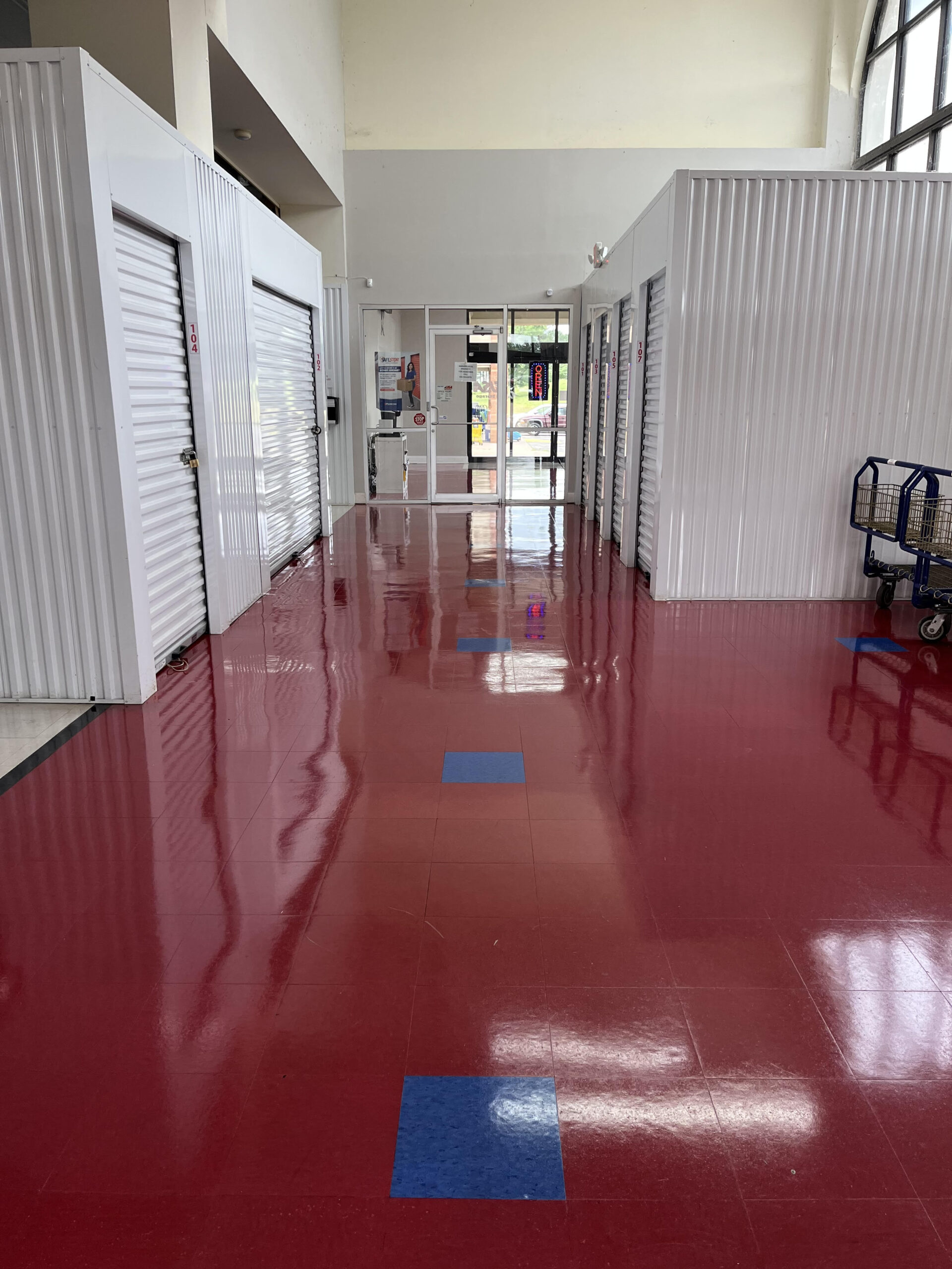 Lonsdale 1 A hallway with shiny red tile flooring, a few blue tiles, white metal storage unit doors on the left, and a cart on the right; glass doors and windows are visible at the end of the hall.
