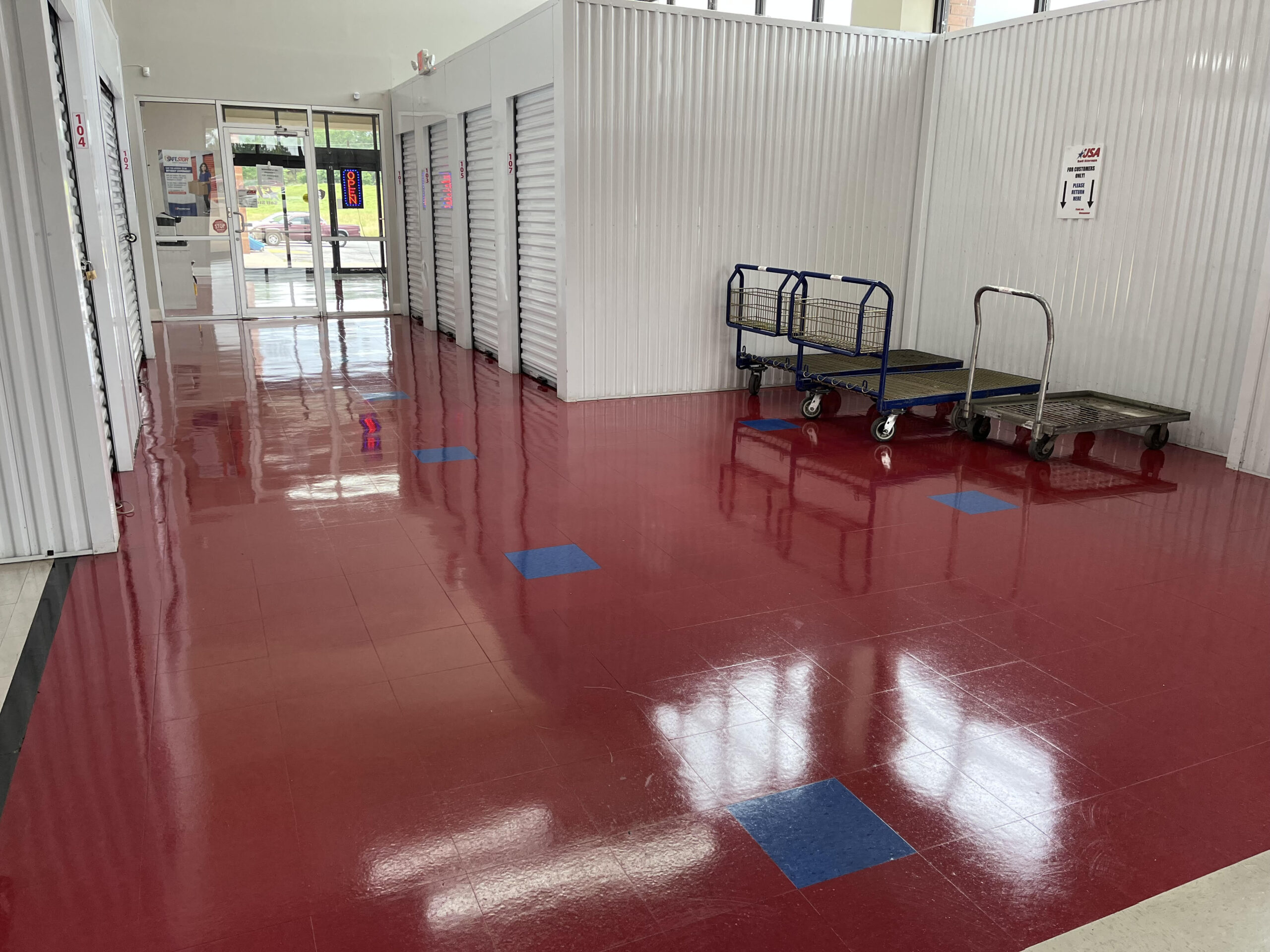 Lonsdale 2 A clean, shiny hallway in a storage facility with red and blue square tiles, metal roll-up doors, and two large platform carts on the right side near the wall.